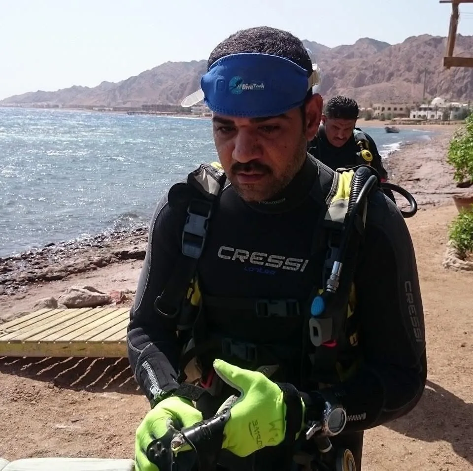 Osama in his wetsuit on the shore at Dahab, checking his regulator, Sinai mountains behind him